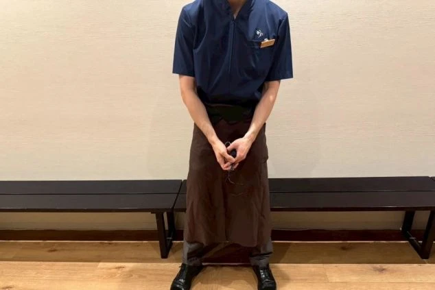 レストラン男性スタッフの制服。紺色のVネックシャツに、濃い茶色のエプロン、灰色のパンツを着用。