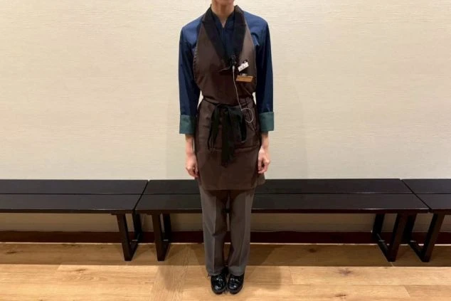 レストラン女性スタッフの制服。紺色の長袖シャツの上に、濃い茶色の和風エプロンを着用し、灰色のパンツを合わせている。