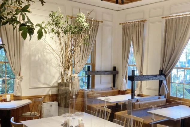 白い壁と木の床、大きな観葉植物が配置され、明るく開放的な空間。テーブル席はパーテーションで区切られており、感染症対策にも配慮している。ホール・レストランサービスのリゾートバイトをアピール。