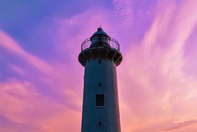 夕焼けに染まる空を背景に、白い灯台がそびえ立っている画像。空はピンク色と紫色が混ざり合い、幻想的な雰囲気を醸し出している。リゾートバイトの休日に観光を楽しめる、風景の画像。