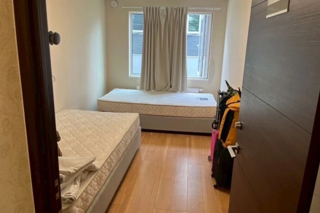 二つのマットレスベッドがある小さな寮の部屋。窓にはカーテンがかかっていて、部屋の隅には旅行用のスーツケースやバッグが置かれている。