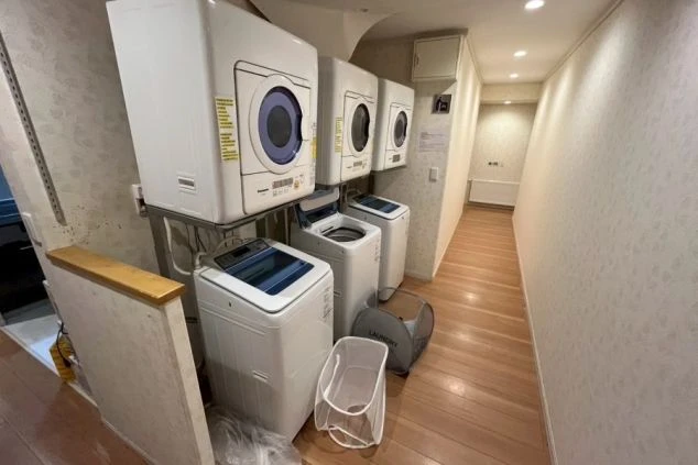 洗濯機と乾燥機が複数台並んでいる共用のランドリーエリア。部屋の床は木製で、壁は花柄の壁紙。