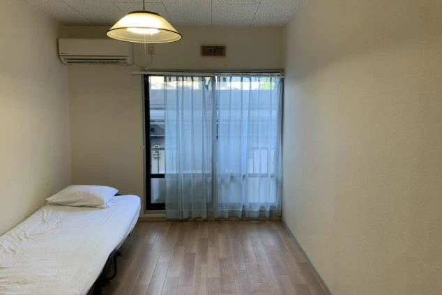 リゾートバイトの寮の部屋の画像。部屋にはベッドが一つ置かれ、フローリングの床と大きな窓が特徴的である。リゾートバイトの寮の部屋の画像。