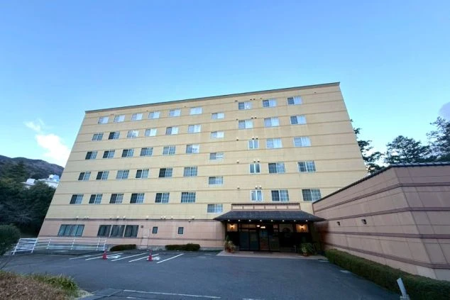 山あいに建つ、クリーム色の多層階ホテル。屋外駐車場とエントランスが見える。