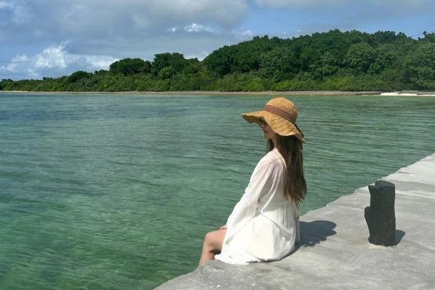  沖縄の桟橋に座り、海を眺める女性。白いワンピースと麦わら帽子がリゾート感を演出し、リゾートバイトで心安らぐ時間を過ごせることをアピール。