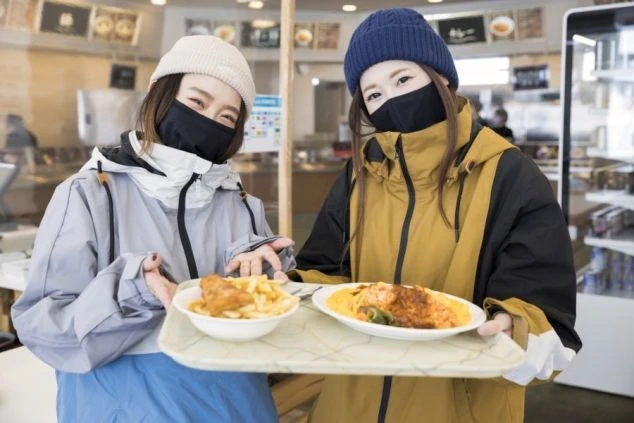 スキー場のレストランで、ランチの乗ったトレーを持って笑顔を見せる女性二人。リゾートバイトでは、こうしたゲレンデ食（ゲレ食）を楽しめるのも魅力の一つです。