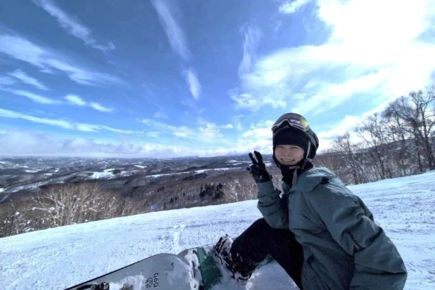 スキー場のゲレンデで、スノーボードに座ってピースサインをする女性。青い空と雲、そして遠くに広がる山々の景色が、冬のリゾートバイトの魅力を伝える。