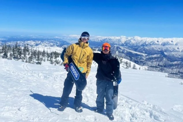 スキー場の山頂でスノーボードを持つ男性2人。快晴の空の下、雪山と雄大な景色が広がっており、リゾートバイトで最高のロケーションを楽しめることをアピール。