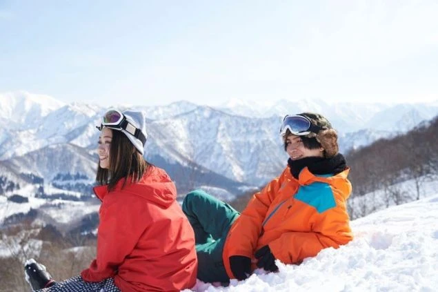 壮大な雪山をバックに、若い男女2人が雪の上に座って休憩している画像。