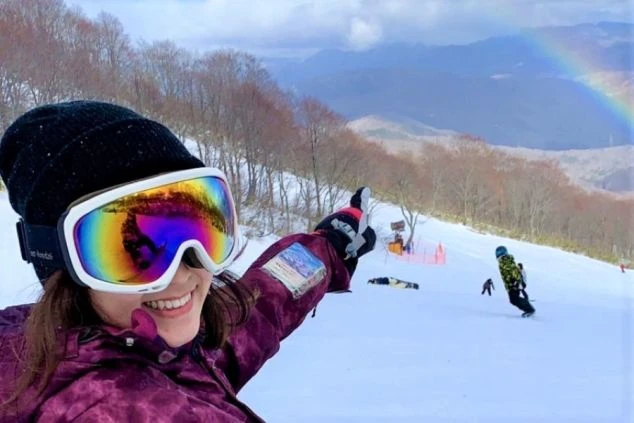 スキー場のゲレンデで、笑顔でポーズをとる女性の画像。遠くに虹が見える。