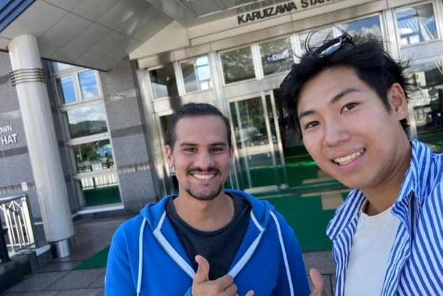 2人の男性が、軽井沢の建物の前で笑顔でカメラに向かってポーズをとっている画像。リゾートバイトの休日の過ごし方や、人間関係の雰囲気を伝えるワクトリスタッフが撮影したオリジナル画像。