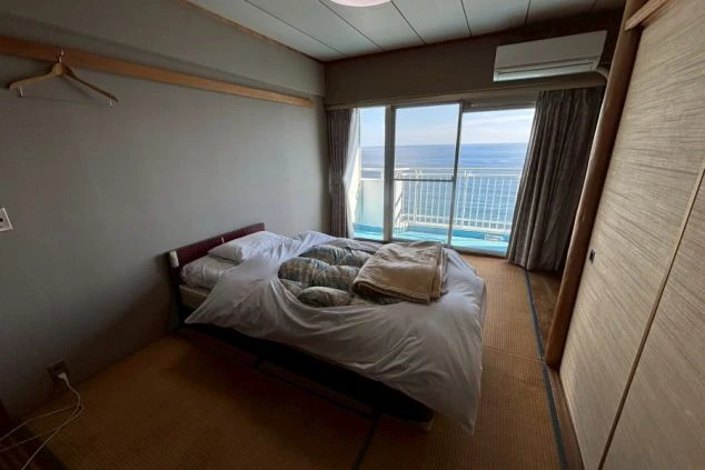 寮の居室。畳敷きの部屋にベッドが置かれ、大きな窓の外には海が見える。部屋にはエアコンとクローゼットが備え付けられている。
