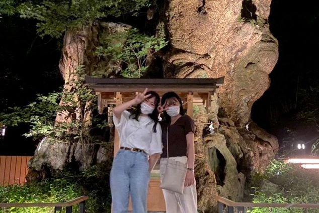 2人の女性ワクトリスタッフが、夜の神社で大きな木を背景に笑顔でピースサインをしている画像。リゾートバイトの休日に観光を楽しんでいる様子が伝わってくる画像。