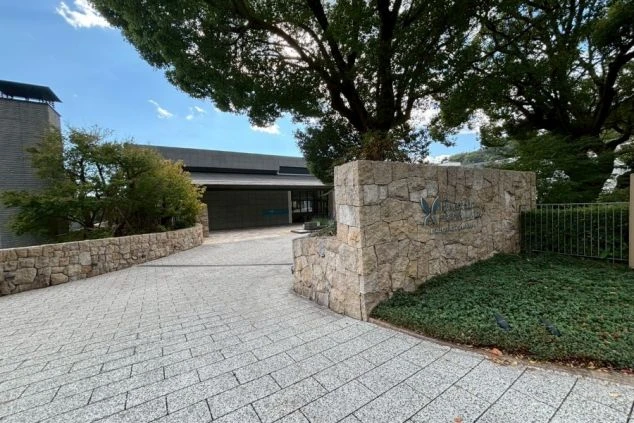 エントランス。積み上げた石垣にホテルのロゴ（蝶のマーク）が刻まれたサインがあり、その奥に低層でモダンなデザインの建物が見える。