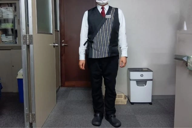 白いシャツに黒のスラックス、和風のデザインが施されたベストを着用した、ホテルの男性フロントスタッフの制服。落ち着いた雰囲気の旅館やホテルでのリゾートバイトを検討している方に魅力的な画像。