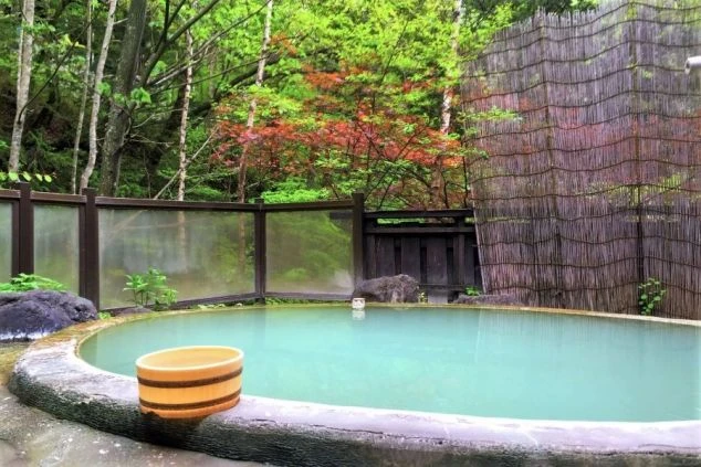 緑豊かな自然の中に設置された露天風呂の画像。浴槽は石で囲まれており、お湯はエメラルドグリーンに輝いている。リゾートバイトの寮やホテルの福利厚生としても利用できる、温泉の画像。