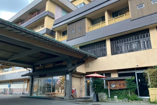 外観。ベージュと茶色の階層的な建物で、手前には車寄せの屋根とマイクロバスの一部が見える。