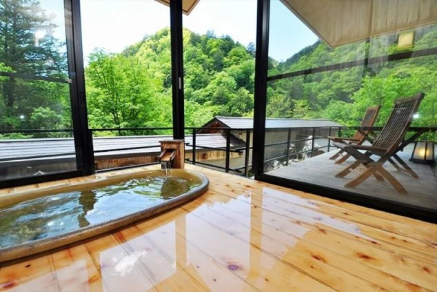 緑豊かな山々が見える、ホテルの温泉の画像。浴槽は木目調で、窓の外にはテラスと椅子が置かれている。リゾートバイトの寮やホテルの福利厚生としても利用できる、温泉の画像。