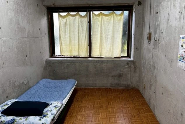 リゾートバイトの寮の個室。コンクリートの壁とフローリングのシンプルな部屋に、ベッドが備え付けられています。