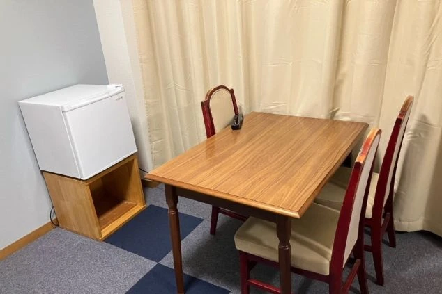 寮の部屋。木製テーブルと椅子、小型冷蔵庫が並ぶスペースが映る。