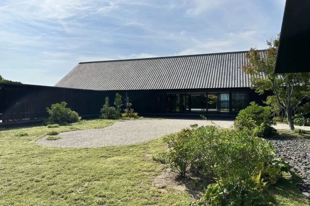 建物の正面。大きな瓦屋根と黒い壁のモダンな和風建築で、全面ガラス戸の向こうに室内が見える。手前は手入れされた芝生で、飛び石がある。