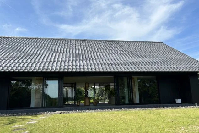 建物の正面。大きな瓦屋根と黒い壁のモダンな和風建築で、全面ガラス戸の向こうに室内が見える。手前は手入れされた芝生で、飛び石がある。