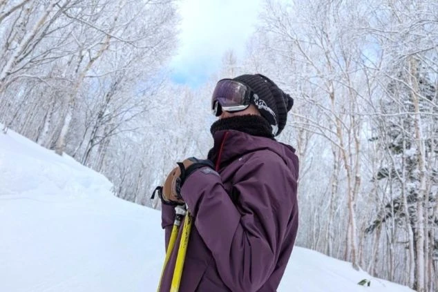 スキー場でスキーウェアとゴーグルを着用した女性。雪化粧した白樺の木々が立ち並ぶゲレンデに立っており、冬のリゾートバイトの魅力を伝える。