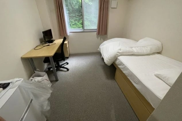 リゾートバイト先の寮の寝室の画像。ベッドの他、デスク・チェア・冷蔵庫がある。