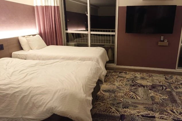 白い寝具が整えられた大きなベッドが中央に置かれたホテルの一室です。部屋の奥には大きな窓があり、外の夜景が見えます。右側にはテレビが壁掛けされ、木目調の壁がアクセントになっています。