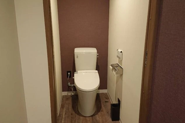 狭い空間にある白いトイレが、茶色の壁と白い壁に挟まれて置かれています。奥の壁は濃い茶色で、手前の壁は薄い色です。