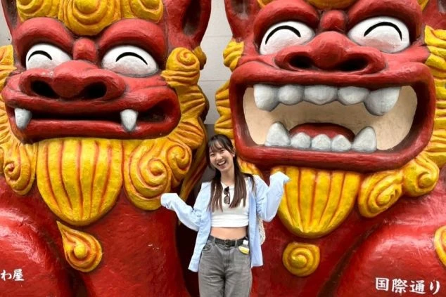 沖縄の国際通りで、巨大なシーサー像の間で笑顔でポーズをとる女性。リゾートバイトの休日に、南国らしい観光地を楽しめることをアピール。