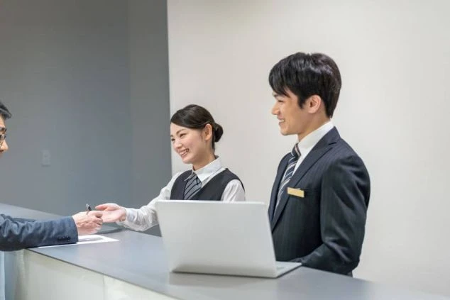 ホテル・旅館のフロントで、笑顔でお客様を迎える男女スタッフ。スーツや制服を着ており、リゾートバイトのフロント業務の雰囲気を伝える。