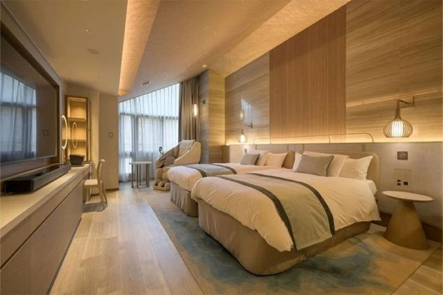 2つのベッドが並べられた、広々としたホテルの客室の画像。部屋にはマッサージチェアやテレビが備え付けられており、窓からは光が差し込む。リゾートバイトの寮やホテルの客室の画像。