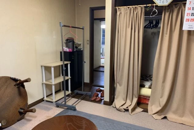 リゾートバイトの男性寮の部屋の画像。テーブルやハンガーラックが置かれ、カーテンで仕切られた収納スペースもあります。