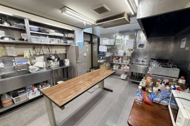 寮の広い共用キッチン。中央に長い作業テーブルがあり、奥にはコンロや冷蔵庫、食器棚が並んでいます。