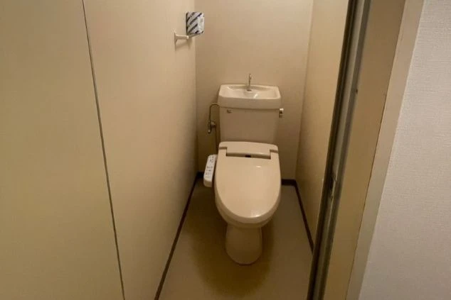 シンプルな内装のモダンな部屋。床は木製で、部屋の奥には窓があり、白いレースのカーテンと黒い遮光カーテンがかけられている。窓の右側にはテレビと小さなテーブル、左側には棚と箱が置かれている。