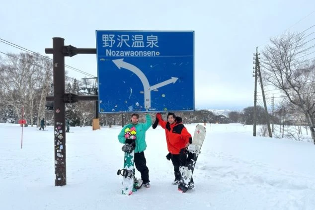 野沢温泉の看板の下でスノーボードを持つ二人。冬のリゾートバイトは、仕事の後にウィンタースポーツを楽しめます。 