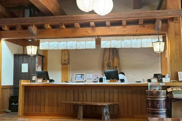 木を基調とした、和風の旅館のフロントカウンターの画像。カウンターにはパソコンが置かれ、スタッフが対応している。リゾートバイトの仕事内容を表す、フロント業務の仕事風景の画像。