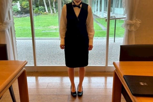 フロントで働く女性スタッフの制服。白いシャツにネイビーのベストとボウタイを着用。窓の向こうには緑豊かな庭が見える。