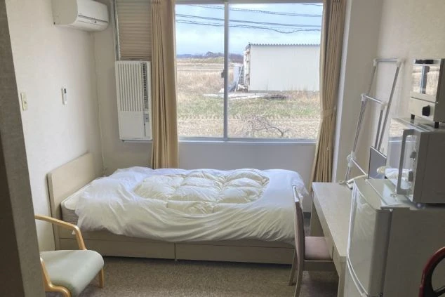 リゾートバイトの個室寮の画像。ベッド、机、冷蔵庫、電子レンジが完備された快適な部屋。窓からはのどかな風景が見えます。