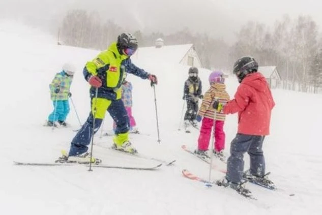 スキーウェアを着用したスキーインストラクターが、雪の中で子供たちにスキーを教えている画像。リゾートバイトの仕事内容を表す画像。