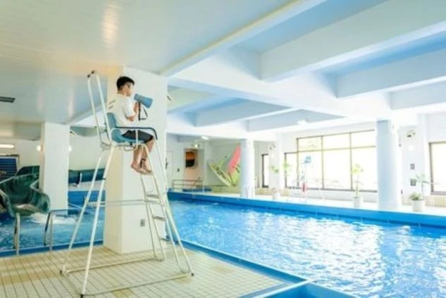 室内プールで監視員のスタッフが監視塔に座り、メガホンを持って注意を促している画像。水色の壁と大きな窓が特徴的で、リゾートバイトの仕事内容を表す画像。