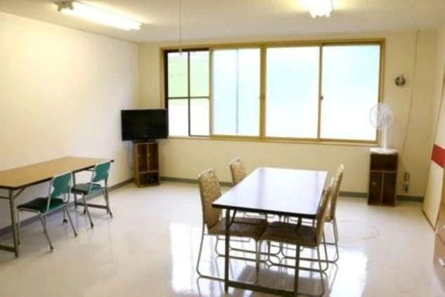 明るい照明と白い壁が特徴的な寮の共同食堂の画像。テーブルと椅子が複数置かれ、テレビや扇風機が備え付けられている。リゾートバイトの食事や休憩に利用できる共有スペースの画像。