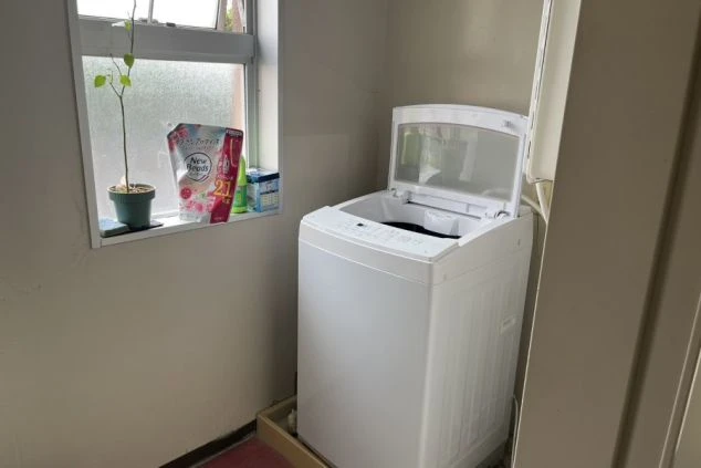 寮の共用スペースに設置された、窓際の縦型洗濯機と洗濯洗剤。