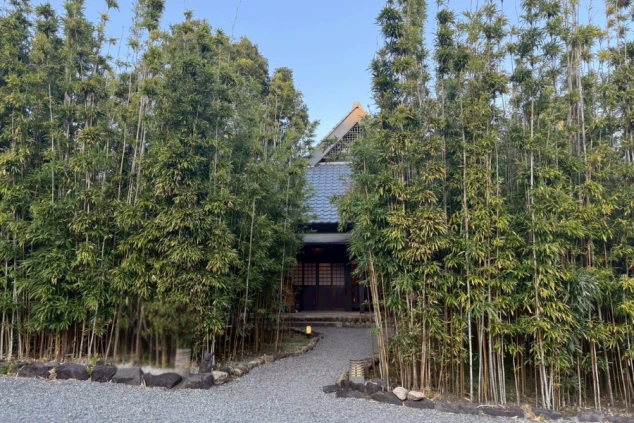 高い竹林に囲まれた小道の先に佇む、伝統的な瓦屋根の和風建築。砂利道が玄関へと続き、落ち着いた静寂な雰囲気が漂っています。