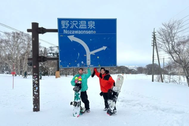 野沢温泉の看板の下でスノーボードを持つ二人。ウィンタースポーツを仕事と両立させたい方に最適です。