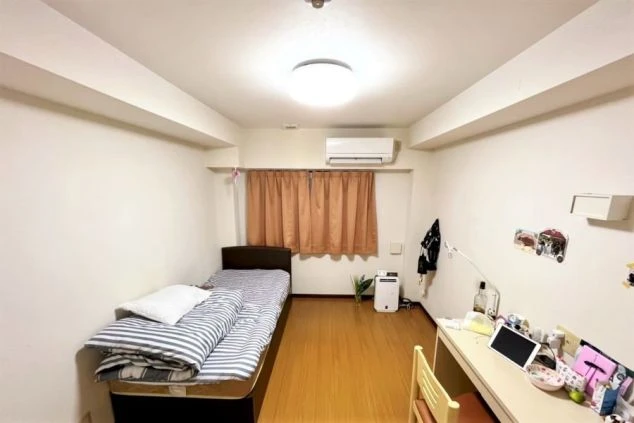 アパートの一室。壁は一面白でフローリングは木目調です。シングルベットが置いてありスタンドライトを置いた白いテーブルがあります。