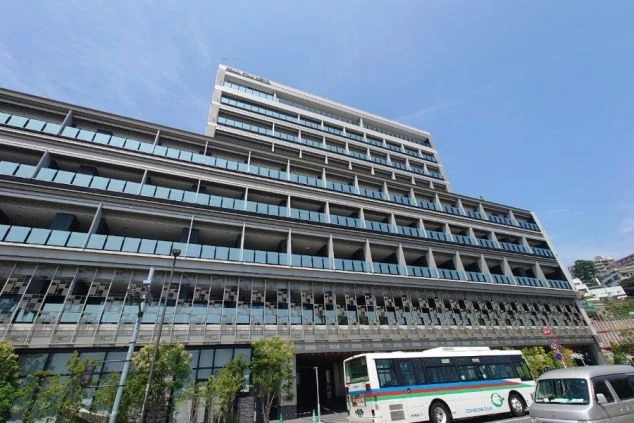 建物全景。ガラス張りのバルコニーが連なるモダンな多層階のホテルで、1階部分には植栽とエントランスが見える。手前には横断歩道と道路が写っている。