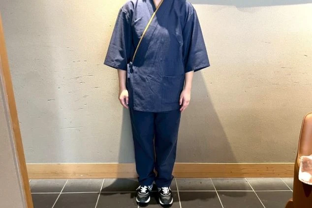 濃紺の作務衣(和風ユニフォーム)のような上下と、黒いスニーカーを着用した人物の全身。薄茶色の壁を背景に正面を向いて立っている。