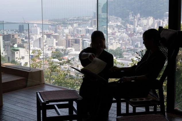 海と街並みが見える窓際で、お客様にサービスを提供する旅館のスタッフ。お客様と向き合うリゾートバイトの接客風景。